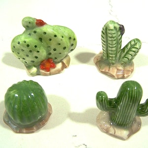 May include: Four ceramic cactus figurines in varying shapes and sizes. The cacti are primarily green with some red and black accents. Each sits on a small, tan base. The figurines are decorative.