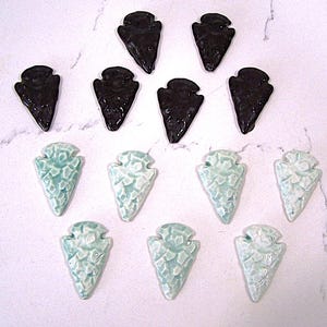 May include: Ten ceramic arrowhead-shaped objects. Six are black and glossy, while the other four are light blue with a textured, cracked appearance. The arrowheads are arranged in two rows on a white surface.