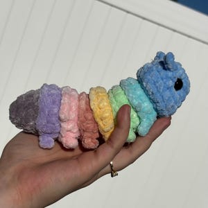 Puede incluir: Un juguete de oruga de peluche hecho a mano en un arcoíris de colores, incluyendo morado, rosa, amarillo y azul. La oruga tiene un cuerpo segmentado y una cabeza pequeña con ojos negros. El juguete es suave y parece estar tejido a ganchillo.