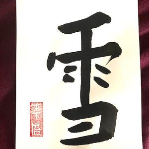 May include: A white rectangular sheet of paper featuring a large black ink calligraphy character for snow. In the lower left corner, there is a small vertical red rectangular stamp containing white Japanese characters.