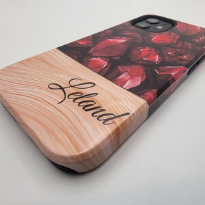 May include: A phone case with a two-tone design. The top half features a pattern of red gemstones, while the bottom half has a wood grain texture. The name "Leland" is written in cursive on the wood grain.