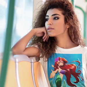Puede incluir: Una mujer con una camiseta blanca con una imagen de dibujos animados de Ariel, la Sirenita, en la parte delantera. Está sentada en un asiento amarillo en un vehículo de transporte público. Lleva pantalones de cuadros rojos y negros.