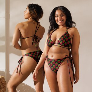 Puede incluir: Dos mujeres con bikinis negros y rojos con estampado de fresas. Los tops de bikini son de forma triangular y las partes de abajo son de lazo lateral.
