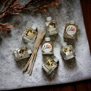 May include: Six small glass bottles containing clear liquid and dried flowers. The bottles have white labels with the text "Lilee Soy and Candle" printed on them. The bottles are arranged on a white fluffy surface with wooden sticks.