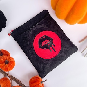 Może przedstawiać: Czarna kosmetyczka z czerwonym okręgiem z czarnym wzorem ust. Kosmetyczka otoczona jest pomarańczowymi dyniami, czarną dynią i szkieletową dłonią. Kosmetyczka ma czerwony zamek błyskawiczny.