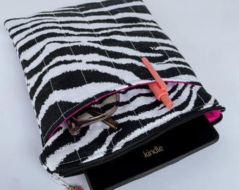 May include: A black and white zebra print quilted case with a pink interior, holding a Kindle, glasses, and a coral pen. The case has a black zipper and a pink heart charm attached.