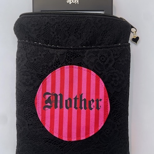 May include: Black lace Kindle sleeve with a pink and red striped circle that says "Mother" in gothic font. The sleeve has a black zipper and a heart charm. A Kindle device is partially visible inside.