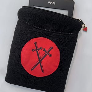 May include: Black lace Kindle sleeve with a red circular patch featuring two crossed swords. The sleeve has a zipper closure with a small heart charm. A Kindle device is partially visible inside the sleeve.
