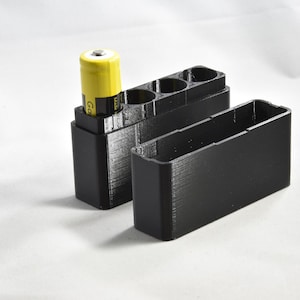 Puede incluir: Una caja de almacenamiento de baterías impresa en 3D de color negro con una batería amarilla en su interior. La caja tiene tres ranuras para baterías y un compartimento separado para guardar baterías.