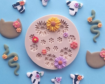 May include: A silicone mold with 16 different shapes, including flowers, cats, snakes, and cows. The mold is pink and has a white background. The mold is filled with colorful polymer clay shapes.