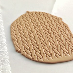 May include: A clear acrylic rolling pin with a raised knit pattern. The rolling pin is used to create a knit texture on a piece of brown clay.