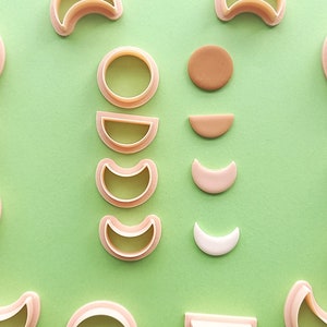 May include: A set of 10 cookie cutters in various shapes, including circles, half moons, and crescent moons. The cutters are arranged on a green surface with small pieces of brown and white clay.