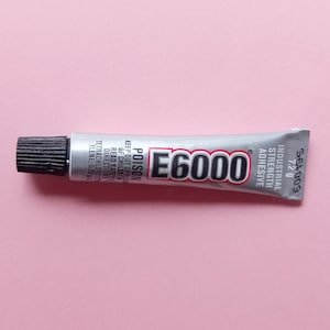 May include: A silver tube of E6000 industrial strength adhesive with a black cap. The tube is labeled with the text "E6000", "POISON", "KEEP OUT OF REACH OF CHILDREN", "READ SAFETY DIRECTIONS", "TETRACHLOROETHYLENE: 0.25", "INDUSTRIAL STRENGTH ADHESIVE", "SEL009", and "7.2 g".