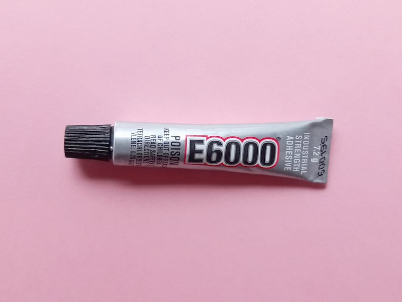 May include: A silver tube of E6000 industrial strength adhesive with a black cap. The tube is labeled with the text "E6000", "POISON", "KEEP OUT OF REACH OF CHILDREN", "READ SAFETY DIRECTIONS", "TETRACHLOROETHYLENE: 0.25", "INDUSTRIAL STRENGTH ADHESIVE", "SEL009", and "7.2 g".