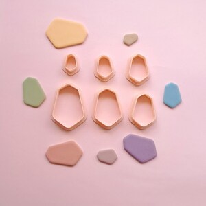 May include: A set of nine plastic cookie cutters in various sizes and shapes. The cutters are in the shape of irregular hexagons with rounded edges. The cutters are arranged on a pink background.