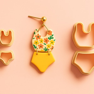 May include: A pair of earrings and two sets of polymer clay cutters in the shape of a swimsuit. The earrings feature a floral design on a cream background, with a yellow bottom. The cutters are light orange.