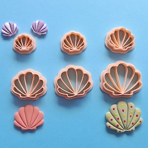 May include: A selection of seashell-themed items, including cookie cutters and decorative shells, displayed on a light blue surface. The cookie cutters are peach-coloured, and the shells are in shades of pink, purple, and yellow with red and yellow dots.
