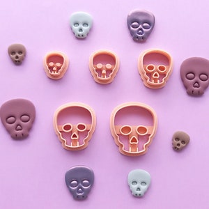 May include: A collection of skull-shaped objects in different sizes and shades, such as light blue, purple, and brown. Some are solid, others are cookie cutters with a peach outline. The items are displayed on a pale purple backdrop.