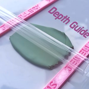 May include: A clear acrylic rolling pin with pink guides labelled "TJ Gallery" on either side. The rolling pin is resting on a piece of green clay. The text "Depth Guide" is printed on the white surface.