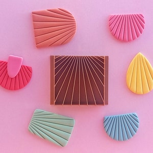 May include: A selection of colourful, geometric clay earrings in various shapes, including half-circles, teardrops, and rectangles. The earrings feature a fan-like design with colours like pink, coral, yellow, and blue. A brown rectangular piece with a sunburst design is also present.