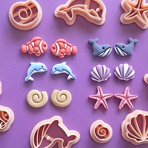 May include: A collection of sea-themed cookie cutters and decorative elements on a purple backdrop. The cutters are shaped like seashells, dolphins, starfish, and fish. The decorative elements include colourful fish, whales, and seashells.