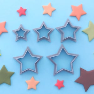 May include: A collection of star-shaped cookie cutters in various sizes, arranged on a light blue background. The cutters are made of metal and have a silver finish. There are also several star-shaped pieces of fondant in various colours, including pink, green, yellow, brown, and orange.