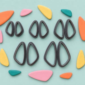 May include: A collection of dark grey teardrop-shaped cutters arranged in two rows, surrounded by various smooth, flat, organic-shaped pieces in orange, pink, yellow, and teal colours, all displayed on a solid light blue background.