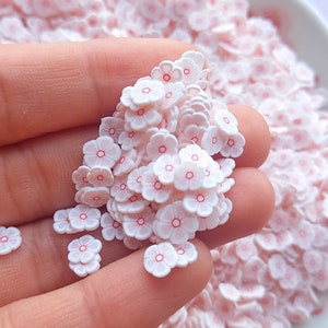 May include: A pile of white flower-shaped sprinkles with pink centers. The sprinkles are small and delicate, perfect for decorating cakes, cupcakes, and other desserts.