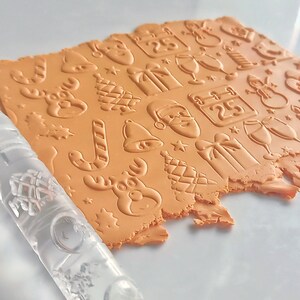 May include: A sheet of orange clay imprinted with Christmas-themed designs, including a reindeer, Santa Claus, gifts, and a calendar. A clear, patterned rolling pin is positioned next to the clay, suggesting its use in creating the embossed pattern.