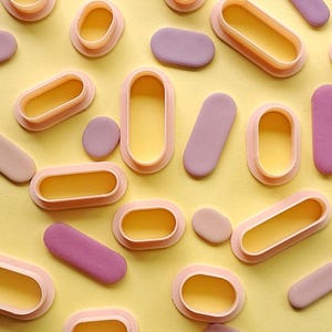 May include: A selection of oval-shaped cookie cutters in various sizes, made from a light pink plastic. The cutters are displayed on a yellow surface.