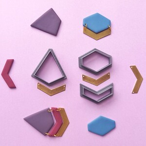 May include: A collection of geometric jewellery components in shades of purple, blue, and gold, arranged on a pink surface. The components include hexagons, triangles, and arrow shapes, suggesting a DIY jewellery-making theme.