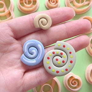 May include: A hand holds three seashell-shaped polymer clay pieces in beige, light blue, and a beige with red and yellow dots. Several seashell-shaped cookie cutters are in the background. The pieces are for crafting or baking.