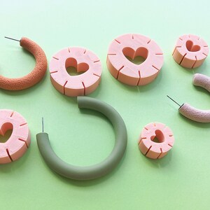 May include: Assortment of colorful handmade earrings featuring heart-shaped cutouts. The earrings are made of polymer clay and come in a variety of colors, including pink, green, and orange.