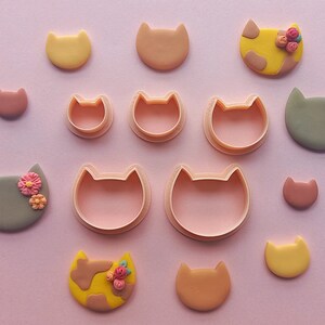May include: A set of 10 cookie cutters in various sizes and shapes, including cats with flowers, plain cats, and hearts. The cutters are made of pink plastic and are arranged on a pink background.
