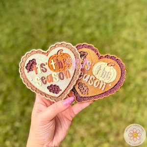 May include: Two heart-shaped ornaments with scalloped edges. The left ornament is cream-colored with an orange pumpkin and the text 'tis the season' in brown. The right ornament is copper-colored with a white pumpkin and the text 'the season' in white.