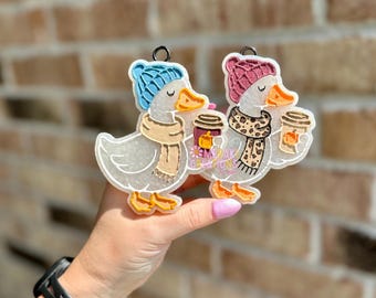 May include: Two duck-shaped ornaments, one with a blue knit cap and the other with a pink knit cap. Both ducks wear scarves and hold coffee cups with pumpkin designs. The ornaments have a fall theme.