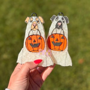 May include: Two Halloween-themed dog ornaments. Each dog is depicted as a ghost holding an orange jack-o'-lantern. The dogs have tan and gray features. The ornaments have a black loop for hanging.