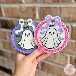 May include: Two circular embroidered patches featuring white ghosts. The left patch has a dark gray background, purple scalloped border, white bow, and candy corn. The right patch has a light background, pink scalloped border, purple bow, and bats.