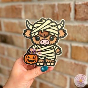 May include: A decorative cutout shaped like a Highland cow dressed as a mummy, featuring cream-colored bandages, a brown coat, and a pink nose. The cow holds a small orange jack-o'-lantern pumpkin. The text Meraki Creations by Payton is visible.