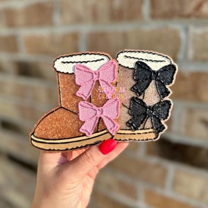 May include: Two decorative brown winter boots with white faux fur trim and bows. One pair has pink bows, the other has black bows. The boots are embellished with glitter and the text "MERAKI CREATIONS".