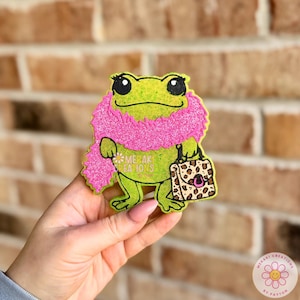 May include: A green frog-shaped accessory with a pink scarf and a leopard print handbag. The frog has large black eyes and the text "Meraki Creations" is visible. The accessory is held against a brick wall background.