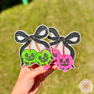 May include: Two embroidered patches featuring glittery black bows attached to pairs of candy apples. One patch shows two green candy apples with jack-o'-lantern faces, and the other shows two pink candy apples with jack-o'-lantern faces.