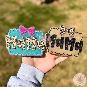May include: Two decorative "Mama" signs. One is teal with a pink bow and leopard print letters. The other is gold with a leopard print bow and black glitter letters. Both signs have a scalloped edge.