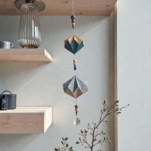 Puede incluir: Móvil colgante geométrico con dos adornos de papel en forma de diamante en verde azulado, dorado y azul y marrón estampados. Cuentas de madera y un cristal cuelgan debajo. El móvil cuelga de una viga de madera.