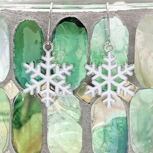 May include: A pair of silver snowflake earrings with white enamel. The earrings are hanging from a glass mosaic background.