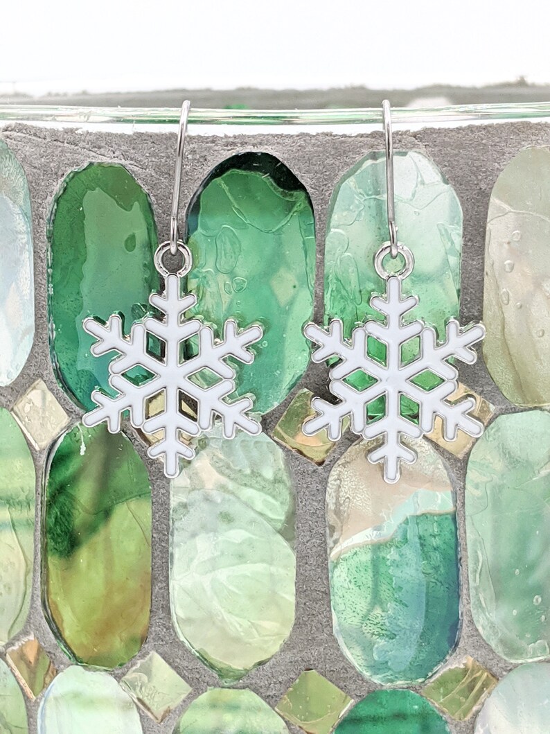 May include: A pair of silver snowflake earrings with white enamel. The earrings are hanging from a glass mosaic background.