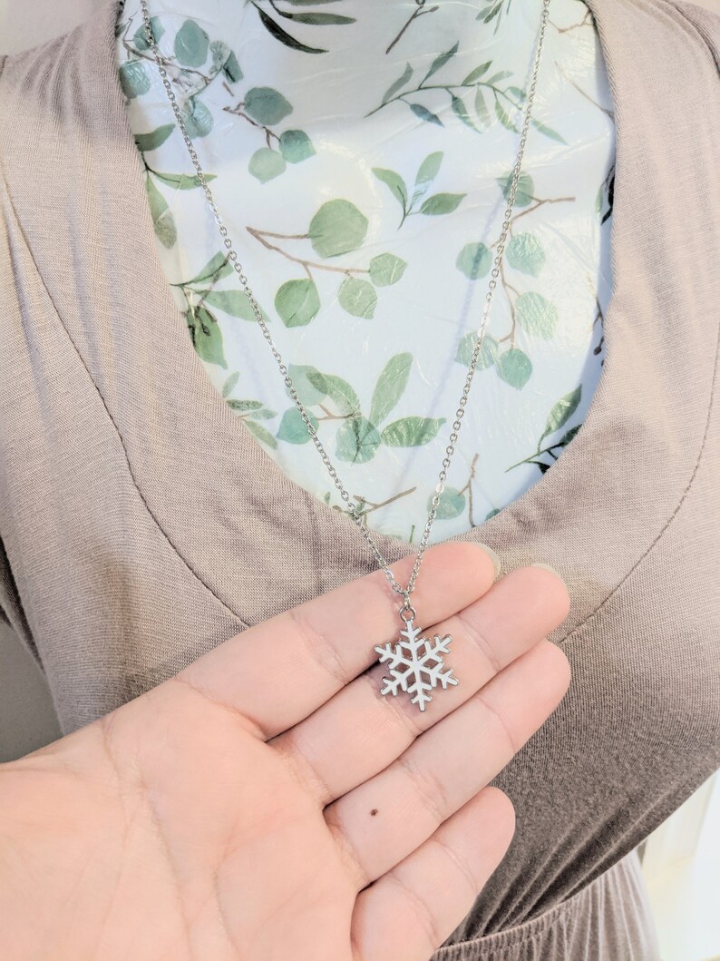 May include: A silver snowflake pendant necklace on a silver chain. The necklace is displayed on a mannequin with a floral pattern.