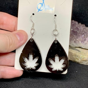 Peut inclure: Une paire de boucles d'oreilles noires en forme de goutte avec une découpe de feuille de marijuana blanche. Les boucles d'oreilles ont des crochets argentés.