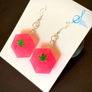 May include: Pink hexagonal resin earrings with a green marijuana leaf design. The earrings have silver hooks.