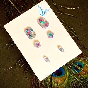 Op de afbeelding: Een set van vijf heldere hars nagelcharms met ingebedde kleurrijke glitter. De charms zijn gevormd als een ster, een traan en twee rechthoeken. De charms worden getoond op een witte kaart met de tekst "Sara & Bella" erop gedrukt.